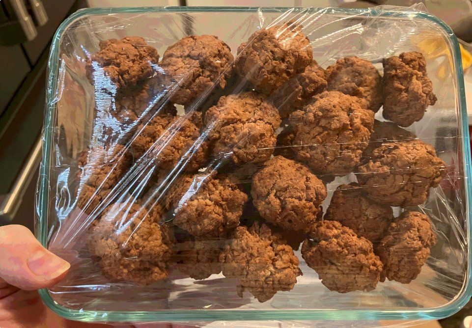 A pile of golden brown coconut macaroons covered by plastic wrap in a square tupperware.